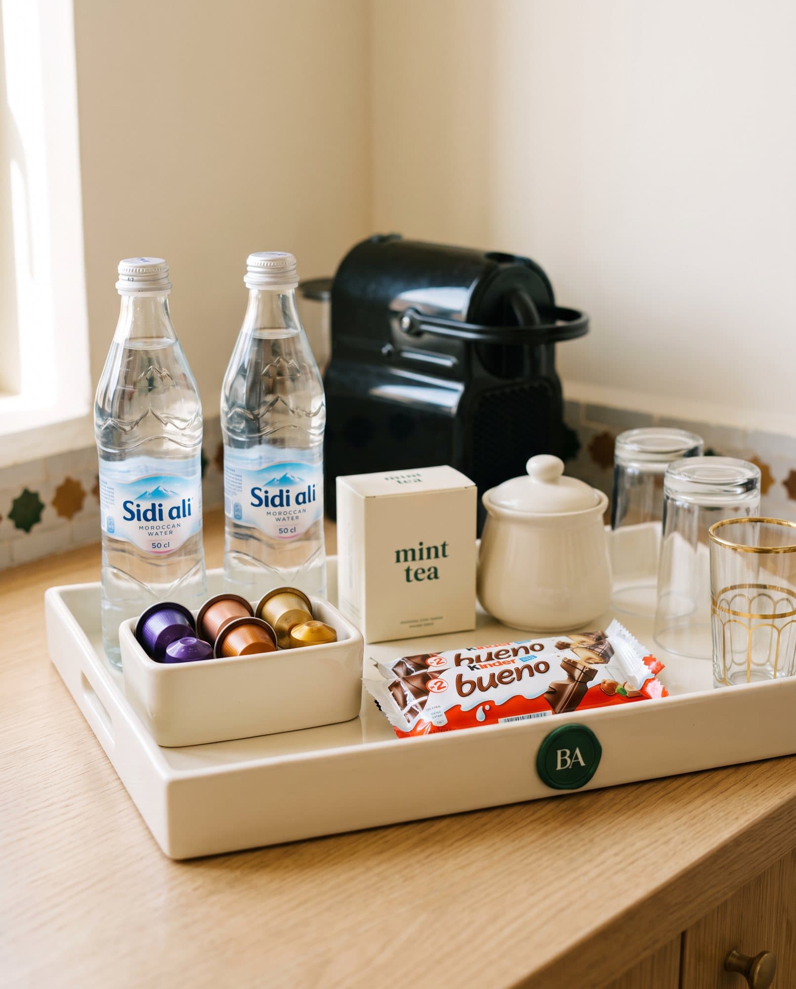 Plateau crème au sceau BA : deux bouteilles Sidi Ali, capsules Nespresso, thé à la menthe, sucrier, Kinder Bueno et verres à thé
