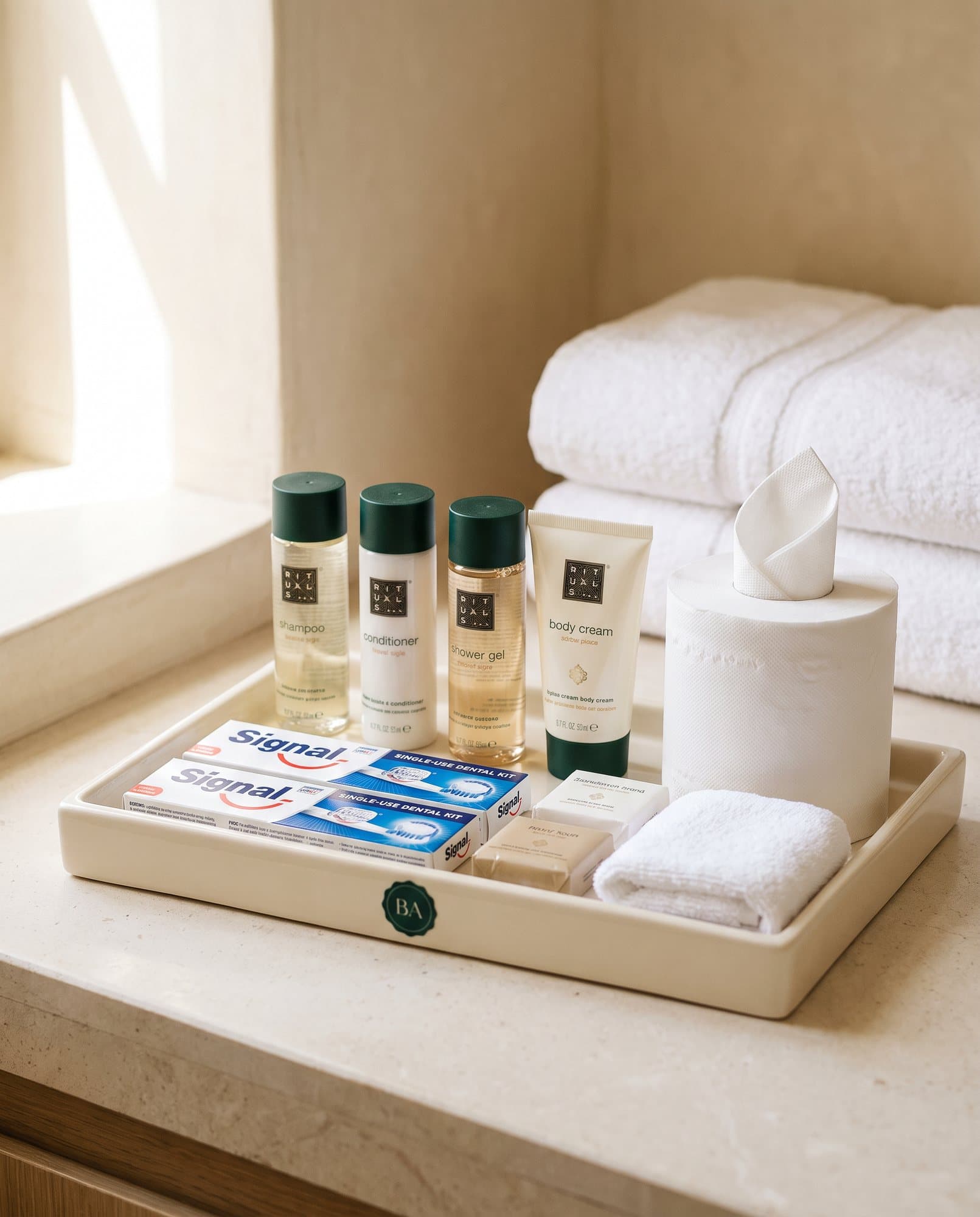 Plateau crème au sceau BA avec minis Rituals, kits dentaires Signal, savons et serviette roulée, dans une salle de bain claire